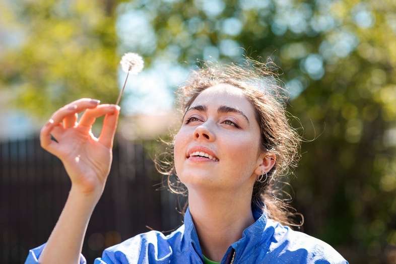 Kvinna i blå jacka håller en maskros och tittar upp mot den utomhus en solig dag.