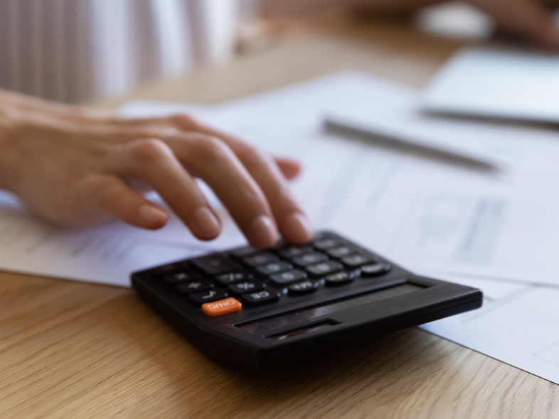 En person använder en miniräknare och arbetar vid en bärbar dator med papper på ett skrivbord.