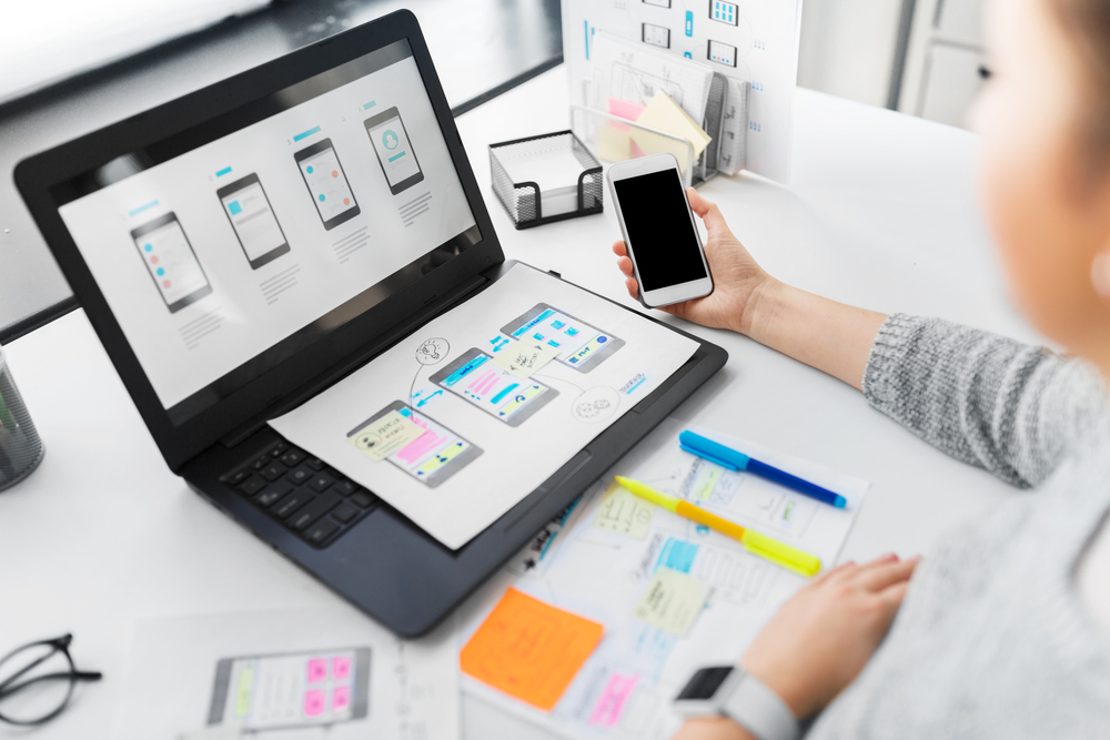 Person arbetar med appdesign på laptop och smartphone vid ett skrivbord med anteckningar och färgpennor.