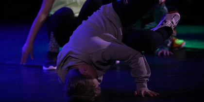 En person utför ett breakdance-move på golvet med stöd av ena handen, andra personer i bakgrunden.