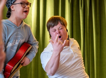Två unga personer, en med gitarr, sjunger och gör rockposer framför gröna gardiner.