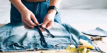 Person arbetar med att sy om ett par jeans, med dragkedja, måttband och sytråd på bordet.