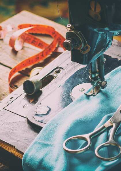 Närbild av en gammal symaskin med blått tyg, sax, fingerborg och måttband på ett träbord.