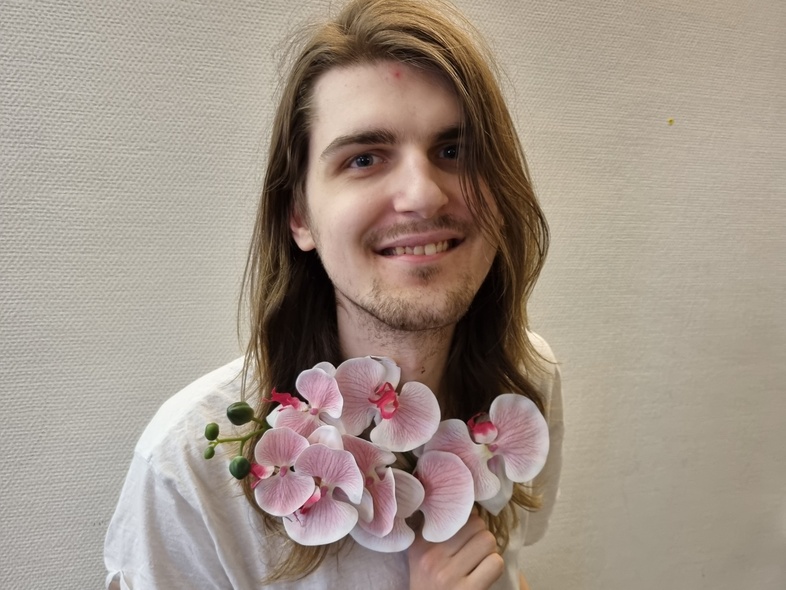 Person med långt hår i vit t-shirt håller en bukett rosa orkidéer framför bröstet, ansiktet är suddat.