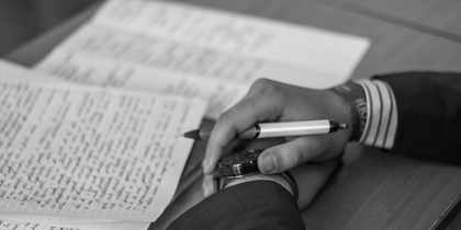 En person i kostym skriver anteckningar för hand på papper vid ett skrivbord, svartvit bild.