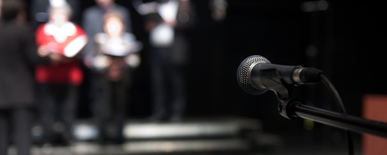 En mikrofon i fokus på en scen med suddiga personer i bakgrunden, troligen en kör eller grupp som förbereder sig.