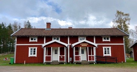 Rött trähus med vita knutar och två ingångar, omgiven av natur och träd under molnig himmel.
