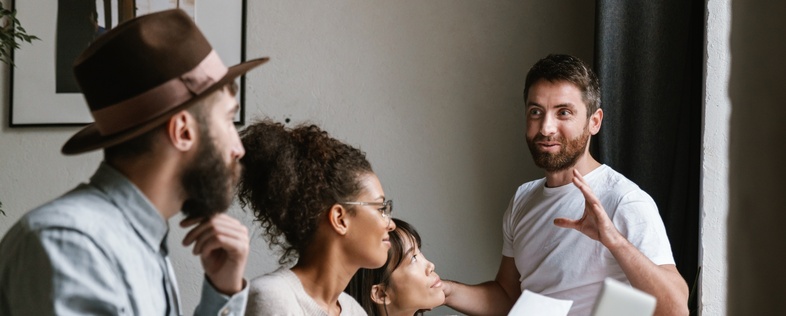 Fyra personer har ett möte vid ett bord, en man står och pratar medan de andra lyssnar.