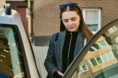 Kvinna med solglasögon öppnar bildörr utomhus vid tegelvägg.