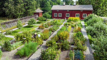 En prydlig trädgård med gångar och odlingsbäddar framför ett rött trähus på landet.