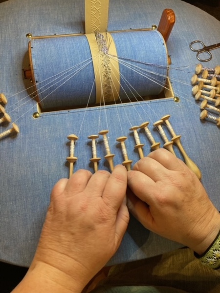 Två händer arbetar med knyppelpinnar på en blå knyppeldyna för att skapa spets.