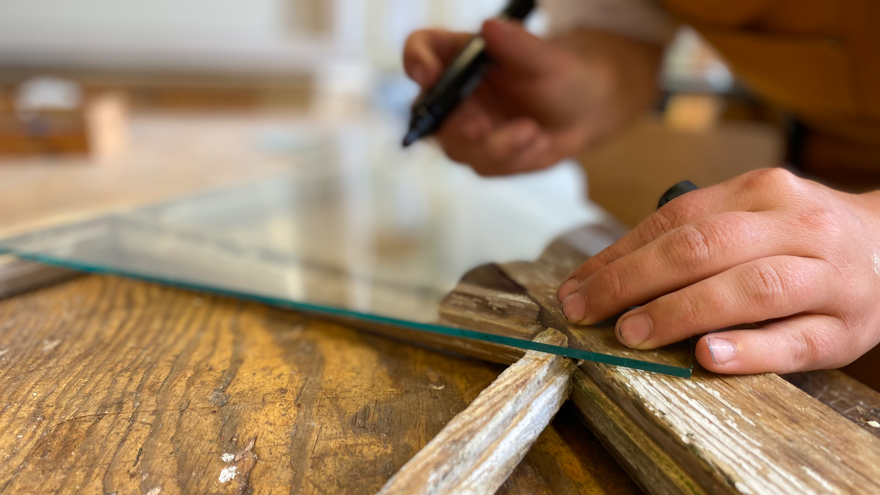 En person markerar en glasskiva med en penna på ett träbord, fokus på händerna.
