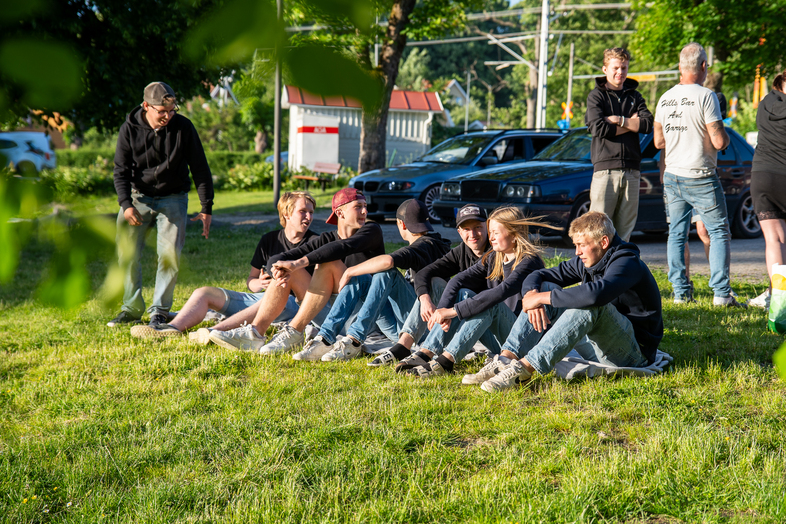 En grupp unga personer sitter på gräset i solen, några bilar och fler personer syns i bakgrunden.