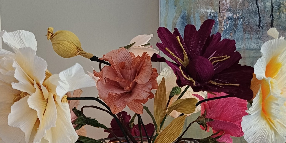 En glasvas med färgglada konstgjorda blommor står på ett turkost bord, med en tavla och dekorationer i bakgrunden.