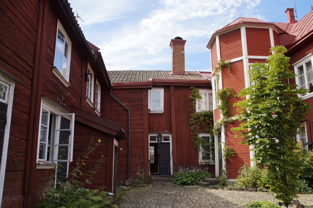 Innergård med röda trähus, kullersten, gröna växter och blå himmel.