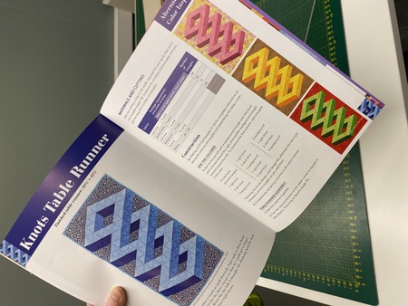 En person håller en öppen bok med instruktioner och bilder på färgglada quiltade bordsdukar i geometriska mönster.