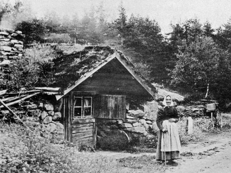En kvinna i traditionella kläder står framför ett litet torp med grästak vid en landsväg, omgiven av skog.