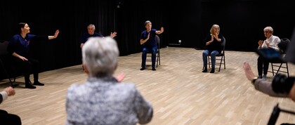 En grupp äldre personer sitter i en cirkel på stolar och gör rörelseövningar i ett ljust rum med trägolv.