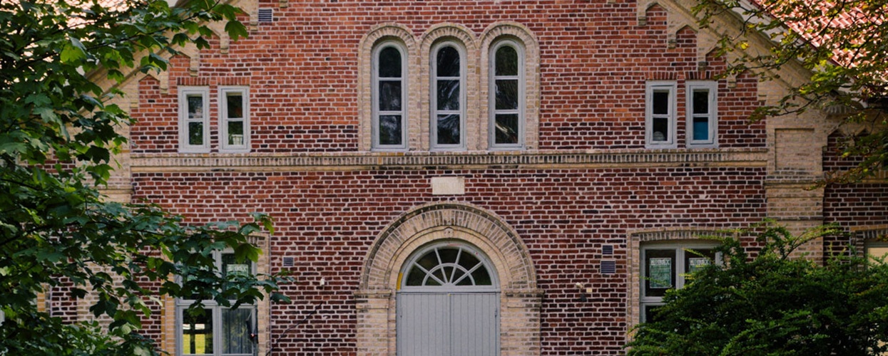 En gammal tegelbyggnad med spetsigt tak, sedd från en träbro omgiven av grönska.