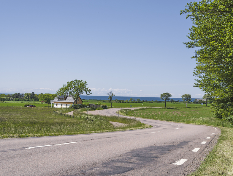 Landsväg som slingrar sig genom gröna fält med ett hus och träd under klar blå himmel.