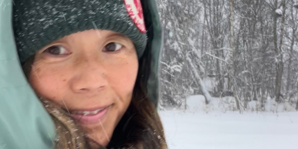 Person i vinterkläder står utomhus i ett snöigt landskap med träd i bakgrunden.