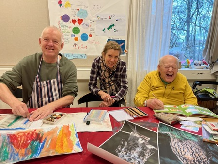 Tre äldre personer sitter vid ett bord och målar tillsammans, glada och engagerade.