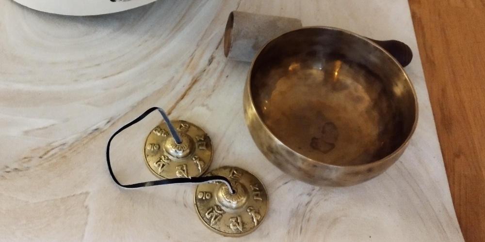 En yogamatta med en vit trumma, klubba, tibetansk klangskål, cymbaler och en röd kudde på ett trägolv.