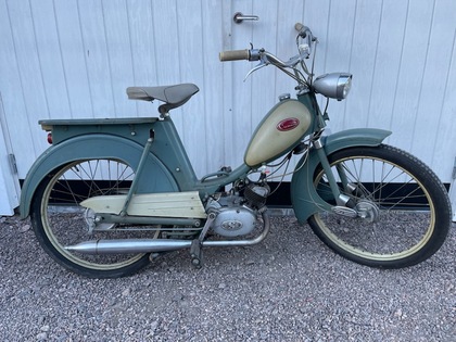 En gammal grön och beige moped står parkerad på grus framför en vit träport.