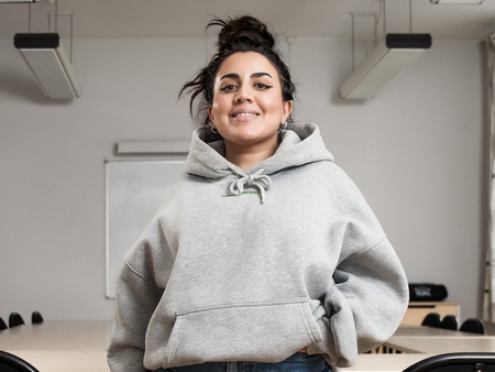Ung kvinna i grå huvtröja står leende i ett tomt klassrum med bord och stolar.