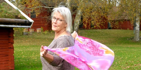 Äldre kvinna med glasögon står utomhus och håller en rosa sjal som blåser i vinden.