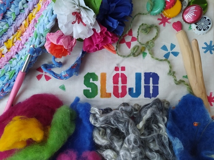 Färgglada slöjdmaterial som garn, tyg, virkverktyg och blommor på en duk med texten 'SLÖJD'.