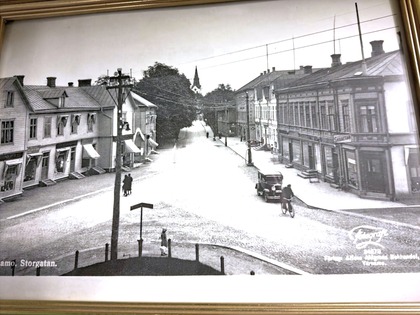 Svartvit bild av en stadsgata med gamla trähus, bil, cyklist och några gående personer.