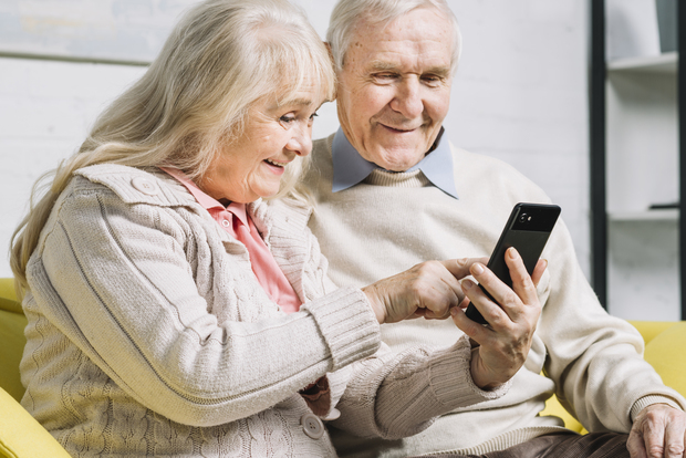 Ett äldre par sitter tillsammans i en soffa och tittar leende på en smartphone.