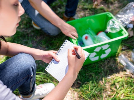 Person skriver i anteckningsblock bredvid återvinningslåda med plastflaskor utomhus.