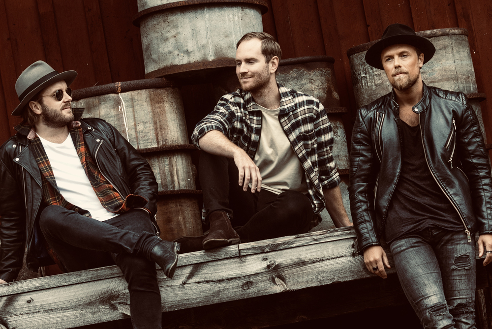 Tre personer sitter och står på en träplattform framför staplade oljefat, klädda i rockiga kläder.