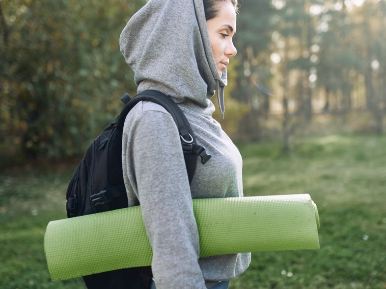 Person i grå hoodie med ryggsäck och yogamatta går utomhus i en park.
