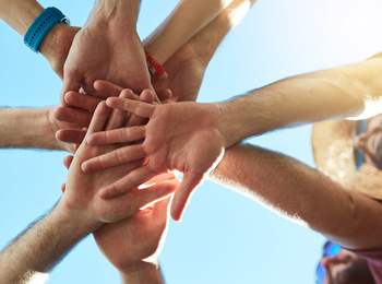Flera personer står i en cirkel och lägger sina händer ovanpå varandra mot en blå himmel.