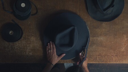En person klipper en mörk hatt på ett träbord, sedd uppifrån. Bandrullar ligger bredvid.