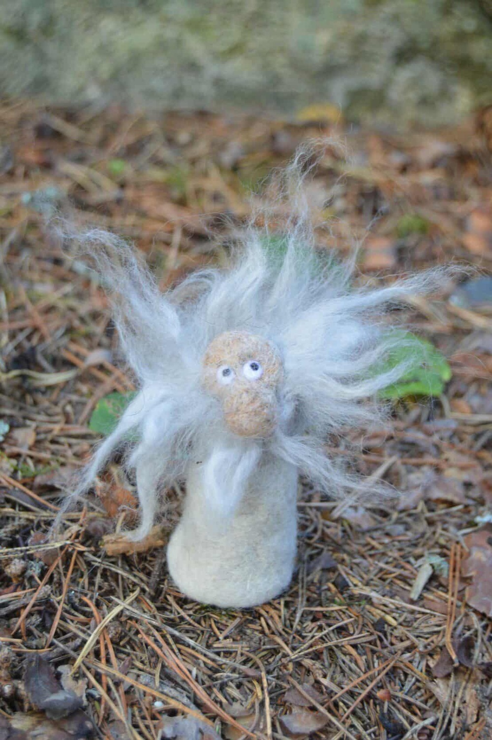 En liten figur av ull med vitt, spretigt hår står på marken i en skogsmiljö.