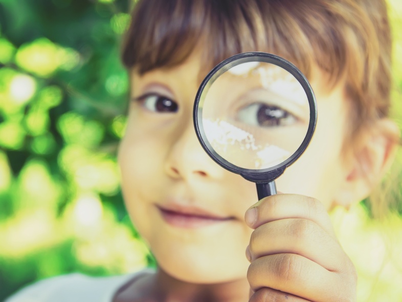 Ett barn håller upp ett förstoringsglas framför sitt öga utomhus, med grönskande bakgrund.