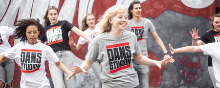 En grupp unga personer dansar energiskt utomhus framför en graffitivägg, alla bär t-shirts med texten 'DANS STUDIO'.