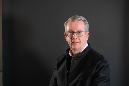 Man med glasögon och grått hår poserar mot en mörk bakgrund, bär svart jacka och vit skjorta.