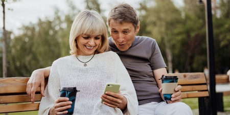 Ett äldre par sitter på en parkbänk, ler och tittar på en mobiltelefon tillsammans.