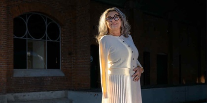 Kvinna i ljus kjol och tröja står på en perrong med tegelvägg och stora fönster i bakgrunden.