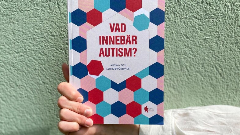 En hand håller i boken Vad innebär autism? mot en grön vägg.