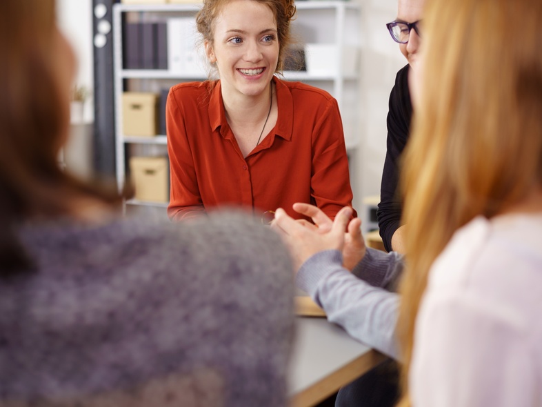 Fyra personer sitter runt ett bord och samtalar, en kvinna i röd skjorta ler mot gruppen.