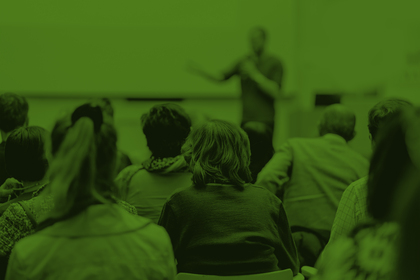 En grupp människor sitter och lyssnar på en föreläsare i ett klassrum, bilden har grön färgton.