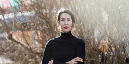 Kvinna i svart polotröja står utomhus med armarna i kors framför torra buskar.