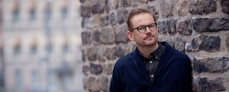 Man med glasögon lutar sig mot en stenmur utomhus, bakgrund med suddiga byggnader.
