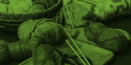 Garnnystan, stickor och sax på en bricka, med en stickad tröja i bakgrunden. Bilden har grön ton.